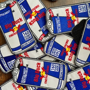 May include: A pile of blue and white embroidered patches shaped like a can of energy drink. The patches have the words "Big Duck" and "Energy Drink" on them. The patches also have the text "160 Flight Hours Before Flight" and "A Little Sassy Gets The Job Done".