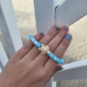 May include: A blue and white beaded bracelet with a white turtle charm. The bracelet is on a person's wrist.