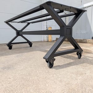 May include: A black metal table base with a geometric design. The base has an X-shaped leg design and is mounted on four caster wheels. The table base is designed to support a tabletop and is suitable for a dining or work table.