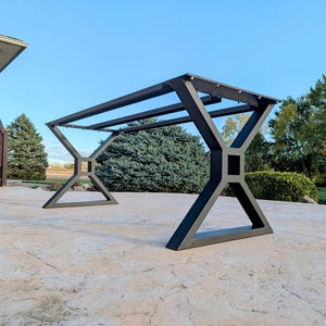 May include: A black metal table frame with an X-shaped leg design. The frame is constructed with geometric shapes and is designed for outdoor use. The table frame is shown on a concrete patio with a blue sky background.