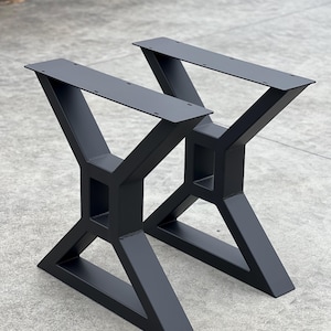 May include: Two black metal table legs with a geometric X-shaped design. The legs are made of thick metal and have a matte black finish.