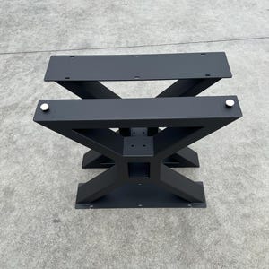 May include: Black metal table base with an X-shaped design. The base has two flat top plates and a lower support structure.