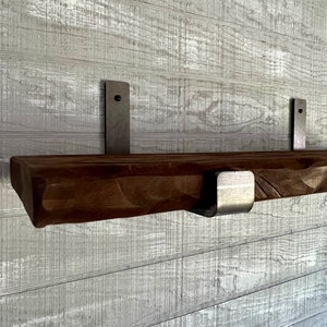 Op de afbeelding: Een houten plank met een rustieke afwerking is bevestigd aan een witgeschilderde muur met behulp van twee metalen beugels. De plank wordt aan beide uiteinden ondersteund door een metalen beugel. De beugels zijn met schroeven aan de muur bevestigd.