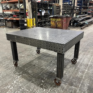 May include: A metal work table with a perforated top and four wheels. The table is made of steel and has a gray finish. The table is in a workshop setting.