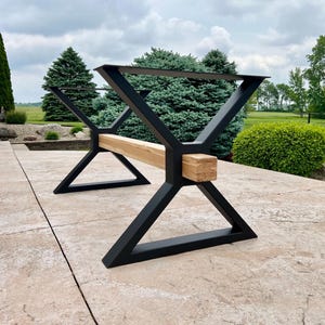 May include: Black metal table base with a natural wood beam. The base has a geometric design with angled legs and a rectangular beam connecting the two sides. The table base is designed for outdoor use.