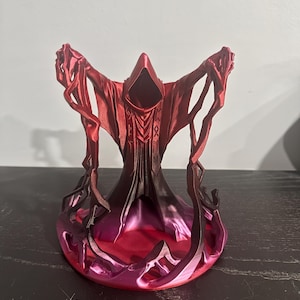 Puede incluir: Escultura impresa en 3D de una figura encapuchada con los brazos extendidos, en tonos rojos y morados. La figura tiene una túnica fluida y está rodeada de llamas estilizadas. La escultura está sobre una superficie oscura.