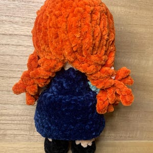 May include: A handmade plush doll with bright orange yarn hair styled in two side braids. The doll wears a dark blue dress, black shoes, and a light blue collar. The doll is made of soft, textured yarn.