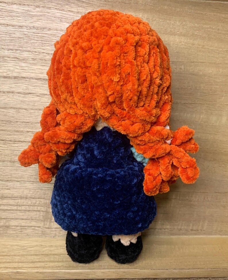 May include: A handmade plush doll with bright orange yarn hair styled in two side braids. The doll wears a dark blue dress, black shoes, and a light blue collar. The doll is made of soft, textured yarn.