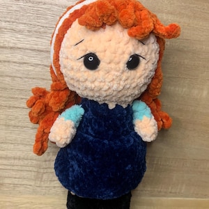May include: A handmade crochet doll with orange hair and a white headband. The doll has large black eyes and is wearing a dark blue dress with light blue sleeves. The doll's feet are black.