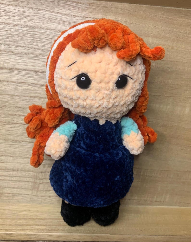 May include: A handmade crochet doll with orange hair and a white headband. The doll has large black eyes and is wearing a dark blue dress with light blue sleeves. The doll's feet are black.