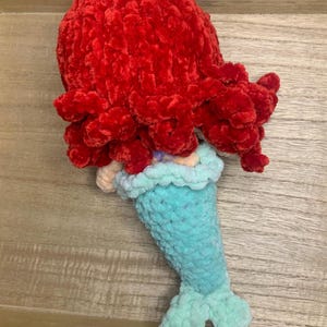 May include: A handmade crochet mermaid doll with a vibrant red head of hair, a light blue tail, and a yellow hair accessory. The doll is crafted from soft, textured yarn, showcasing a whimsical design. The mermaid's tail is approximately 6 inches long.