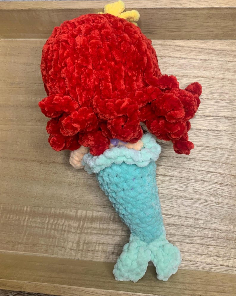 May include: A handmade crochet mermaid doll with a vibrant red head of hair, a light blue tail, and a yellow hair accessory. The doll is crafted from soft, textured yarn, showcasing a whimsical design. The mermaid's tail is approximately 6 inches long.