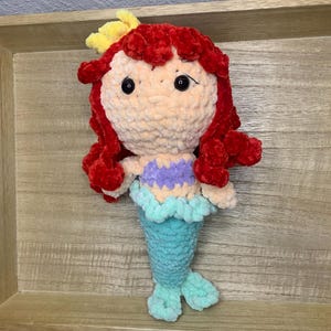 May include: A handmade crochet mermaid doll with red hair, a yellow hair accessory, and a light purple top. The doll has a turquoise tail and fins, and black safety eyes. The doll is displayed on a wooden surface.