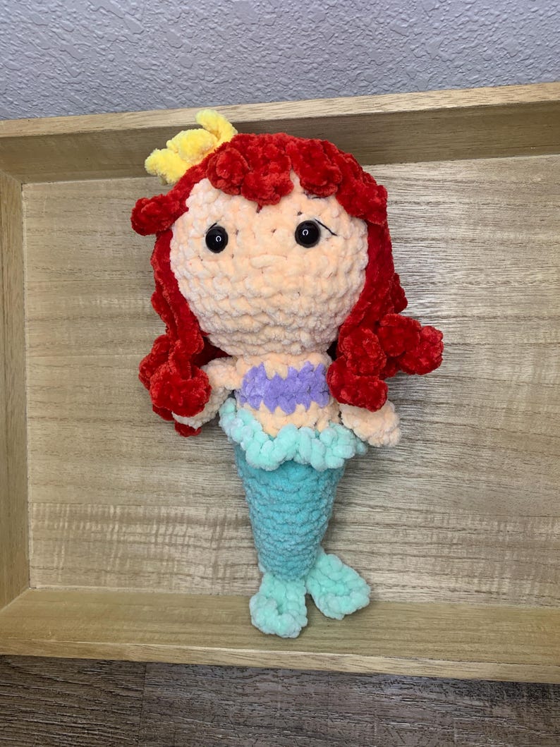May include: A handmade crochet mermaid doll with red hair, a yellow hair accessory, and a light purple top. The doll has a turquoise tail and fins, and black safety eyes. The doll is displayed on a wooden surface.