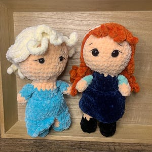 May include: Two handmade crochet dolls, one in light blue and the other in navy blue. The dolls have yarn hair and are displayed on a wooden tray. The dolls have black button eyes and are wearing outfits.