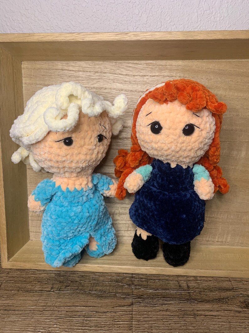 May include: Two handmade crochet dolls, one in light blue and the other in navy blue. The dolls have yarn hair and are displayed on a wooden tray. The dolls have black button eyes and are wearing outfits.