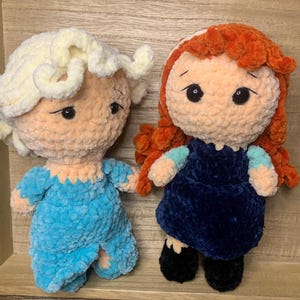 May include: Two handmade crochet dolls. One doll has light blonde hair and a blue outfit. The other doll has red hair and a dark blue dress. Both dolls have large black eyes and are made of soft, textured yarn.