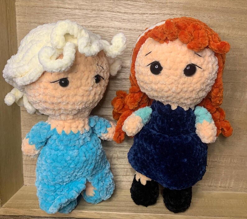 May include: Two handmade crochet dolls. One doll has light blonde hair and a blue outfit. The other doll has red hair and a dark blue dress. Both dolls have large black eyes and are made of soft, textured yarn.