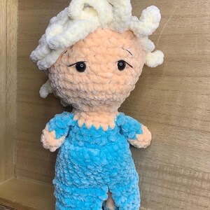 May include: A handmade plush doll with a light peach face and white yarn hair. The doll wears a blue outfit with a scalloped neckline. The doll has black eyes and is made of soft, textured yarn.