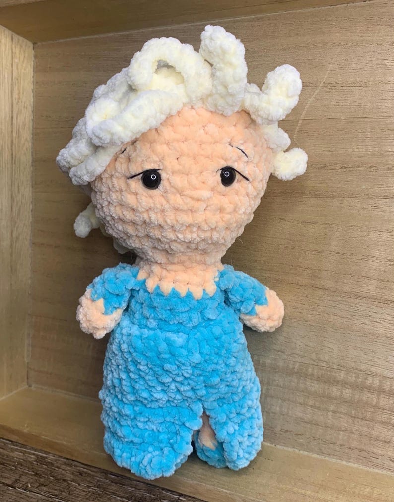May include: A handmade plush doll with a light peach face and white yarn hair. The doll wears a blue outfit with a scalloped neckline. The doll has black eyes and is made of soft, textured yarn.