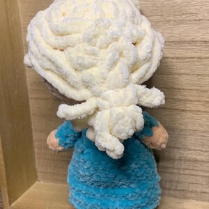 May include: A handmade doll with a fluffy, cream-colored head and a blue dress. The doll's hair is styled in a voluminous, textured design. The doll's arms are a light peach color. The doll is made of soft, plush material.