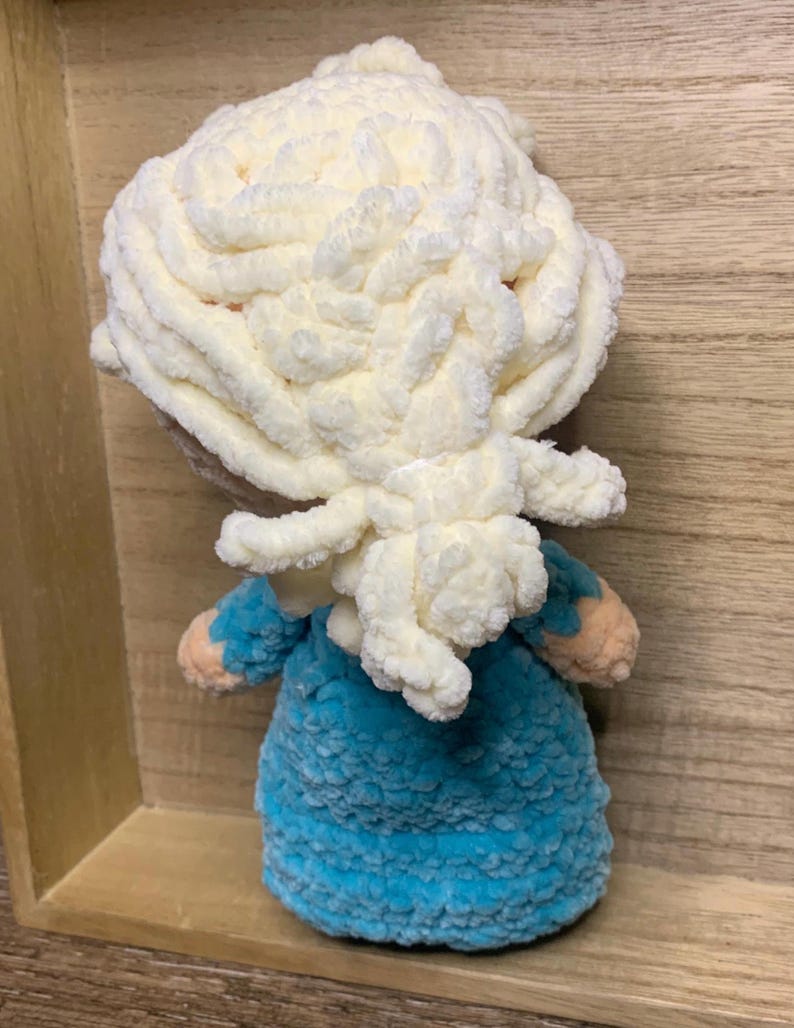 May include: A handmade doll with a fluffy, cream-colored head and a blue dress. The doll's hair is styled in a voluminous, textured design. The doll's arms are a light peach color. The doll is made of soft, plush material.