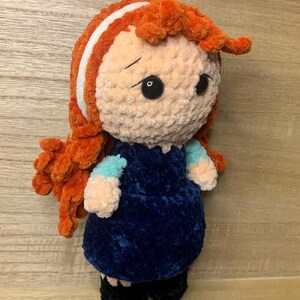 May include: A handmade plush doll with orange hair, a white stripe, and a blue dress. The doll has black eyes and is wearing black shoes. The doll is made of soft, textured yarn.
