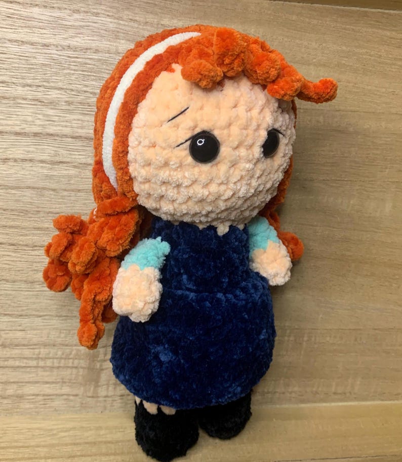 May include: A handmade plush doll with orange hair, a white stripe, and a blue dress. The doll has black eyes and is wearing black shoes. The doll is made of soft, textured yarn.