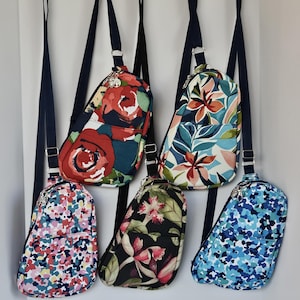 May include: Five different patterned crossbody bags hanging on hooks. The bags are all made of fabric and have a triangular shape. The patterns include floral, abstract, and geometric designs.