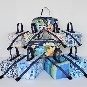 May include: A collection of nine rectangular fabric storage bags with top handles and zippered closures. Each bag features a unique pattern, including floral, geometric, and abstract designs in various shades of blue, green, and multi-colored prints.