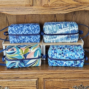 May include: Four blue and white patterned zippered pouches with navy blue handles. The pouches are stacked on a wooden shelf.