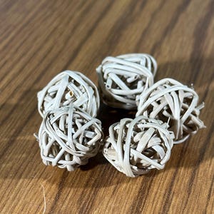 May include: Five small, woven, spherical objects made of light-colored, natural material. The spheres are arranged in a loose cluster on a wooden surface, with visible texture and grain. The woven design creates a textured, open pattern.