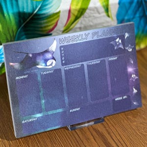 May include: A weekly planner with a galaxy-themed design featuring a manta ray illustration. The planner has spaces for notes, Monday through Sunday, and a section for the week of.