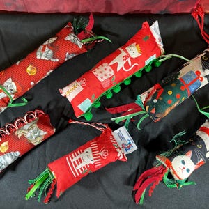 May include: Six cylindrical fabric cat toys featuring various holiday-themed prints of cats, gift boxes, and festive patterns. Each toy includes attached ribbons, pom-poms, or decorative trim, and a small white product tag is attached to each item.