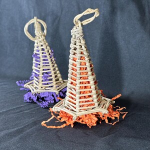 May include: Two cone-shaped woven wicker structures resembling hats, each featuring a loop at the top. One structure is filled with purple crinkle-cut paper, and the other is filled with orange crinkle-cut paper, both resting on a dark surface.
