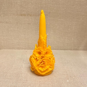 Puede incluir: Una vela amarilla con un diseño floral en la base. La vela tiene una forma alta y cónica y está decorada con pétalos de flores tallados. La vela es de un color amarillo brillante y se encuentra sobre un fondo neutro.