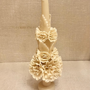 May include: Ivory-colored candle with carved floral and leaf details. The candle features a base, a central column, and decorative layers of carved flowers. The candle is a decorative item, suitable for home decor or special occasions.