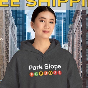 Op de afbeelding: Donkergrijze hoodie met de tekst "Park Slope" en de letters F, G, R, B, Q, 2 en 3 in een ontwerp in metro-stijl. De hoodie heeft een capuchon met trekkoord en een voorzak. Op de achtergrond een stadsstraat.