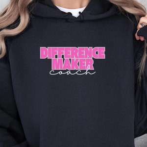 Könnte beinhalten: Schwarzer Kapuzenpullover mit dem Text "Difference Maker Coach" in rosa Schrift.