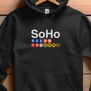 May include: Black hooded sweatshirt with the word "SoHo" in white, above a colorful subway line design. The sweatshirt has a front pocket and a drawstring hood. The garment is displayed on a wooden hanger against a wood-grain background.