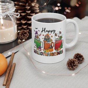 May include: White ceramic mug with a black liquid inside. The mug features a colorful graphic with the word "Happy" and the phrase "Hallowthanksmas". The graphic includes images of gnomes, pumpkins, and other fall-themed elements.