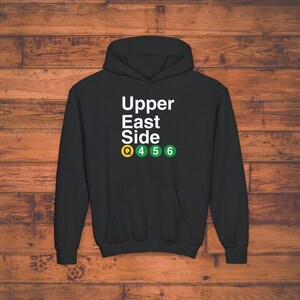 以下が含まれることがあります： 「Upper East Side」という白い文字と、その下に「Q 4 5 6」という文字が書かれた緑と黄色の地下鉄の丸いマークが並んだ黒いフード付きスウェットシャツです。