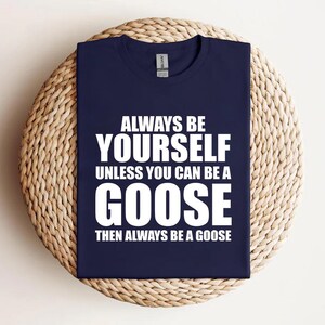 Puede incluir: Camiseta azul marino con el texto blanco: "ALWAYS BE YOURSELF UNLESS YOU CAN BE A GOOSE THEN ALWAYS BE A GOOSE". La camiseta está sobre una superficie redonda y tejida.