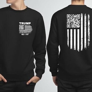 Puede incluir: Sudadera negra con un diseño blanco de la bandera estadounidense y un código QR. La parte delantera muestra "TRUMP" encima de la bandera y "45 * 47" debajo. La parte trasera presenta un diseño de bandera desgastada. Prenda unisex.