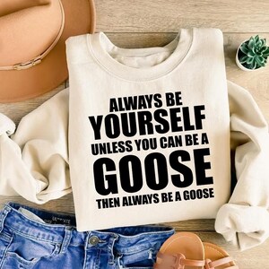 Puede incluir: Sudadera de color crema con el texto "ALWAYS BE YOURSELF UNLESS YOU CAN BE A GOOSE THEN ALWAYS BE A GOOSE" en negro. La sudadera se combina con pantalones cortos de mezclilla azules, un sombrero marrón y sandalias.