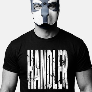 Puede incluir: Una persona con una máscara azul y blanca con orejas de perro y una camiseta negra. La camiseta tiene la palabra "HANDLER" impresa en letras blancas grandes. La máscara cubre la parte superior de la cara.