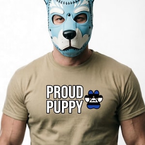 Puede incluir: Una máscara de perro de cuero azul claro con detalles y costuras blancas. La persona lleva una camiseta beige con las palabras "PROUD PUPPY" y un gráfico de huella de pata.
