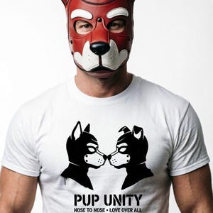 Puede incluir: Una máscara de perro de cuero rojo y blanco con nariz y orejas negras. La persona lleva una camiseta blanca con un gráfico negro de dos perros tocándose la nariz, y el texto "PUP UNITY" con la frase "NOSE TO NOSE • LOVE OVER ALL".