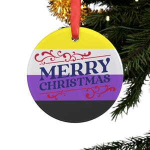 Könnte beinhalten: Ein runder Weihnachtsanhänger aus Keramik mit horizontalen Streifen in Gelb, Weiß, Lila und Schwarz. Der Text MERRY CHRISTMAS ist in Dunkelblau in der Mitte aufgedruckt und wird von dekorativen roten Schnörkeln über und unter dem Text betont.