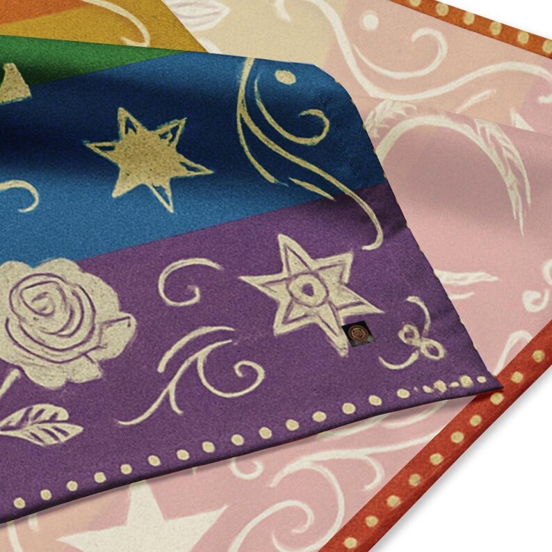 May include: Close-up of a folded fabric with a celestial design. The fabric features a blue section with gold stars, a purple section with a rose and star, and a pink section with a floral pattern. The edges are bordered with red and white dots.